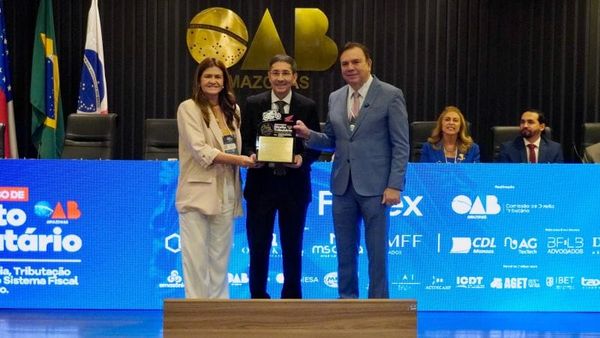 Mulher e dois homens no palco, um deles recebendo uma placa de homenagem da OAB Amazonas, com o logo da OAB ao fundo