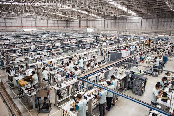 Vista aérea de uma fábrica com dezenas de trabalhadores em suas estações de trabalho, realizando tarefas de montagem