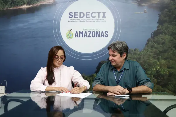 Luzia Raquel Said assina documento ao lado de um homem, com o logo da Sedecti e do Governo do Amazonas ao fundo, em Manaus