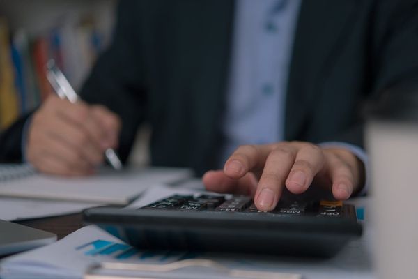 Mãos de uma pessoa em terno digitando em uma calculadora, enquanto a outra mão escreve em um caderno, sobre uma mesa