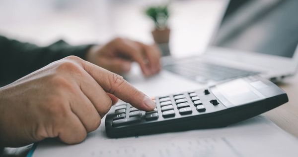 Pessoa utiliza calculadora sobre mesa de escritório com notebook ao fundo.