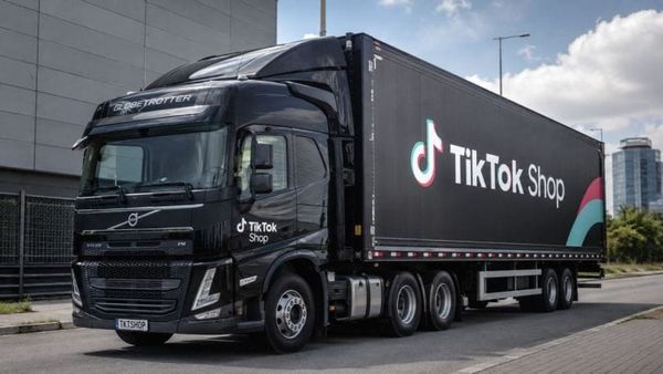 Caminhão preto da Volvo com a marca TikTok Shop na lateral do baú e na cabine, estacionado em frente a um prédio cinza