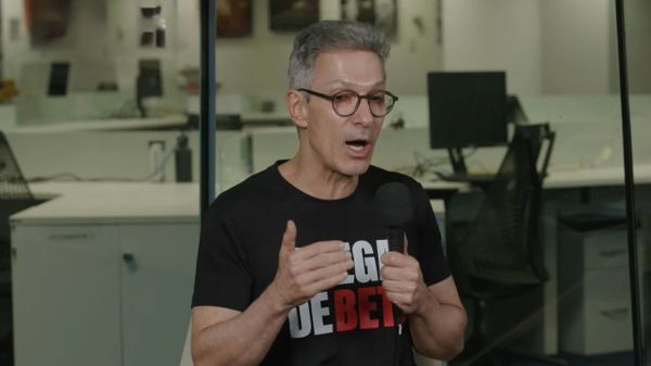 Romeu Zema fala ao microfone durante entrevista em um escritório, vestindo camiseta preta com inscrições em branco e vermelho.