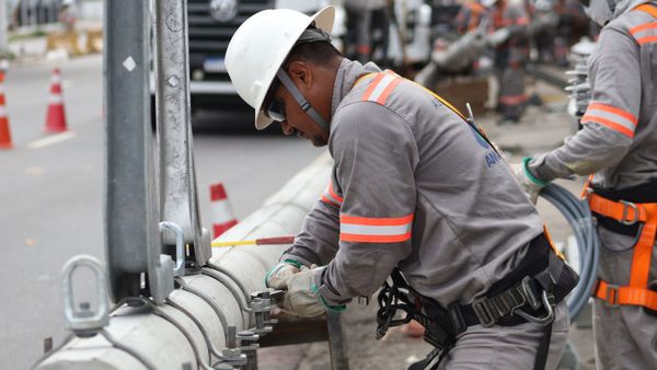Eletricista com capacete branco e uniforme cinza realizando manutenção em poste de energia no Amazonas.