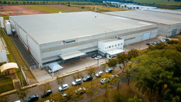 Vista aérea do galpão industrial da ADATA em Manaus, com fachada cinza, logotipo da empresa e carros estacionados à frente.