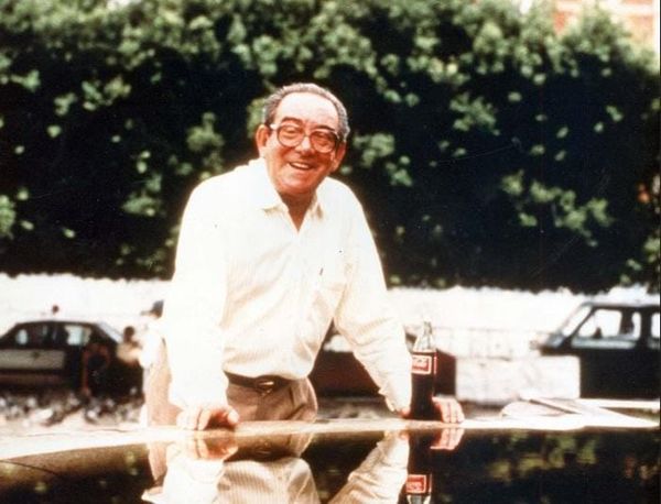 O empresário Antônio Simões sorrindo, usando óculos e camisa clara, apoiado em uma superfície com uma garrafa de Coca-Cola.