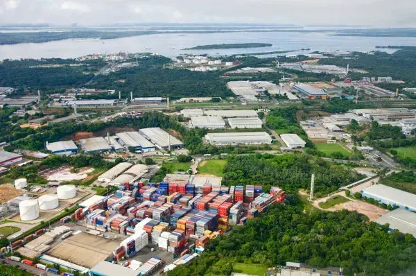 Vista aérea do Polo Industrial de Manaus com galpões de fábricas, contêineres coloridos e o Rio Amazonas ao fundo.