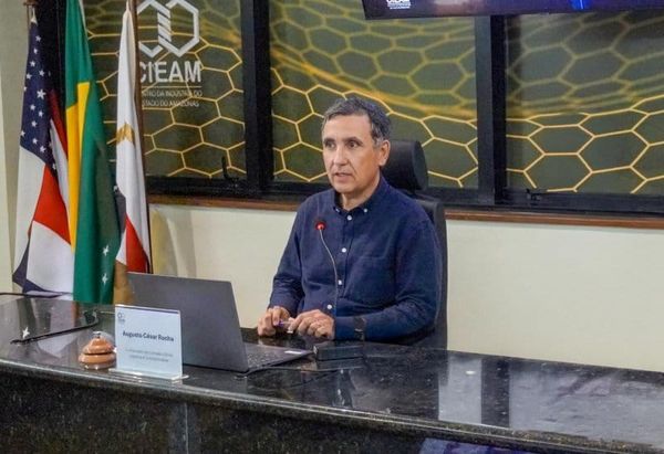 Augusto César Barreto Rocha sentado à mesa com notebook, microfone e bandeiras ao fundo em evento no Cieam, em Manaus.
