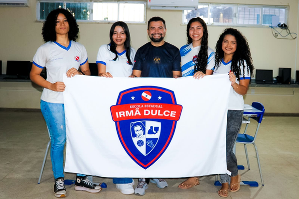 Quatro estudantes e um professor seguram a bandeira da Escola Estadual Irmã Dulce em uma sala de aula com computadores.