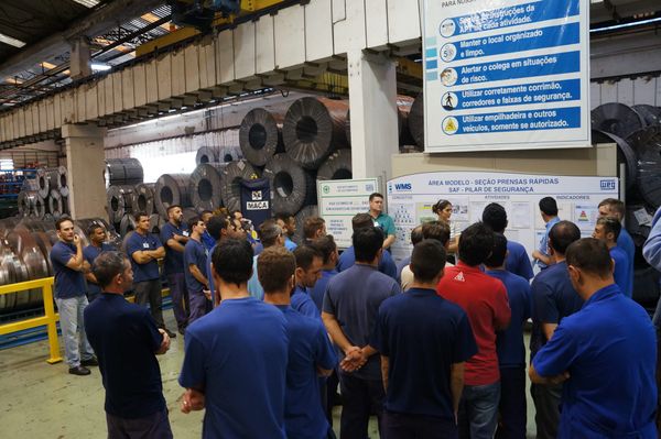 Colaboradores da WEG reunidos em frente a um quadro de gerenciamento de rotina em ambiente fabril com bobinas de aço ao fundo.