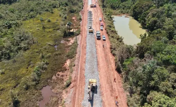 Vista aérea de máquinas e caminhões trabalhando em trecho de terra da rodovia BR-319 cercada por vegetação e água.