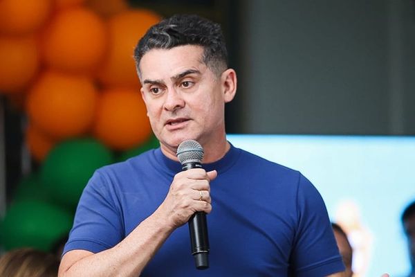 David Almeida fala ao microfone durante evento, vestindo camiseta azul, com balões laranjas e verdes ao fundo.