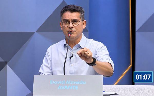 David Almeida fala ao microfone durante debate eleitoral, vestindo camisa azul clara e óculos.