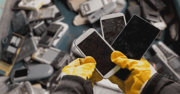 Mãos com luvas amarelas segurando três smartphones com telas quebradas sobre uma pilha de sucatas eletrônicas.