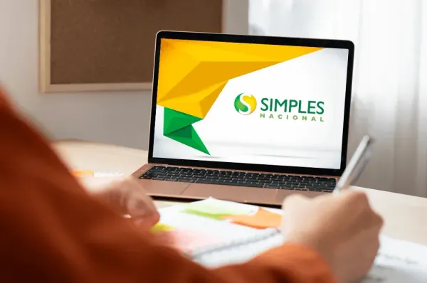 Notebook aberto exibindo a logomarca do Simples Nacional na tela, com uma pessoa escrevendo em um caderno ao lado.
