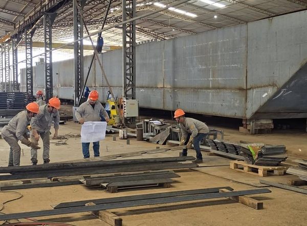 Trabalhadores com capacetes e uniformes manuseiam peças de metal dentro de um galpão industrial no Estaleiro Juruá.