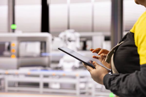 Pessoa com uniforme industrial segurando um tablet em frente a máquinas de uma linha de produção automatizada.