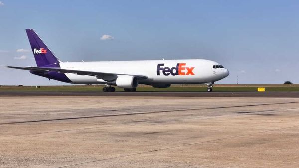 Avião de carga branco com a logomarca roxa e laranja da FedEx estacionado na pista de um aeroporto sob céu azul.