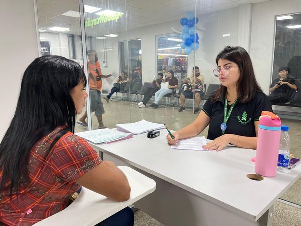 Mulher com camisa de RH entrevista candidata em mesa branca; ao fundo, pessoas aguardam atendimento em sala com vidro.