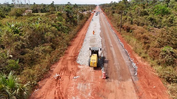 Rolo compressor amarelo trabalha em trecho de terra e brita da rodovia BR-319 cercada por vegetação amazônica.