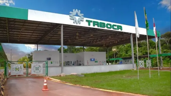 Fachada da entrada da Mineração Taboca com guarita branca, cobertura metálica e bandeiras do Brasil e do Amazonas ao lado.