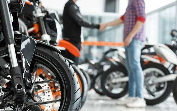 Pneu dianteiro de motocicleta em destaque em uma concessionária com dois homens apertando as mãos ao fundo.