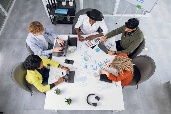 Cinco pessoas sentadas ao redor de uma mesa branca em reunião, analisando gráficos e usando notebooks e tablets.