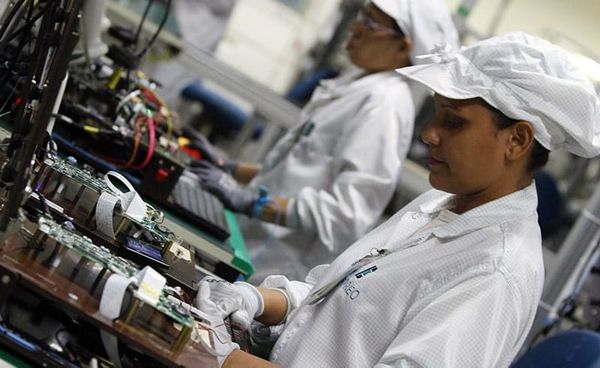 Duas operadoras com uniformes brancos e toucas trabalham em linha de montagem de componentes eletrônicos em fábrica de Manaus.