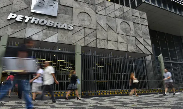 Pessoas caminham em frente ao edifício sede da Petrobras, com letreiro da empresa em destaque na fachada de concreto.