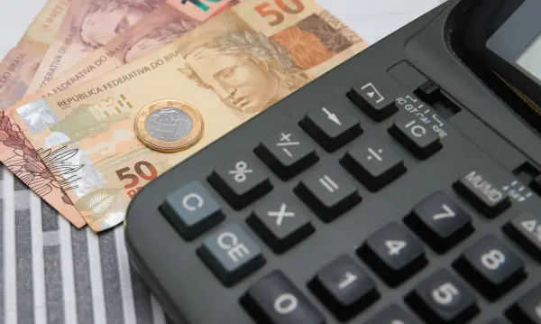 Calculadora preta em primeiro plano ao lado de cédulas de 50 reais e uma moeda de um real.