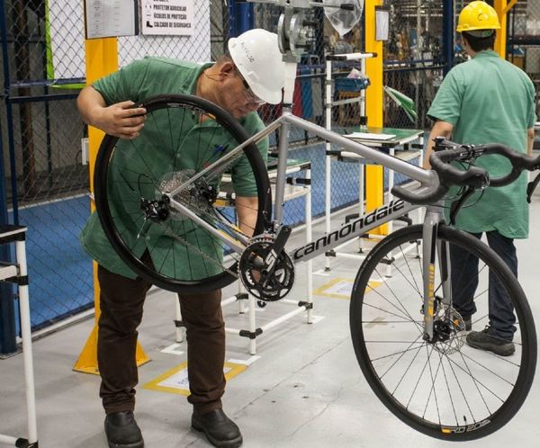 Trabalhador com capacete branco ajusta roda traseira de uma bicicleta prateada da marca Cannondale em fábrica de Manaus.