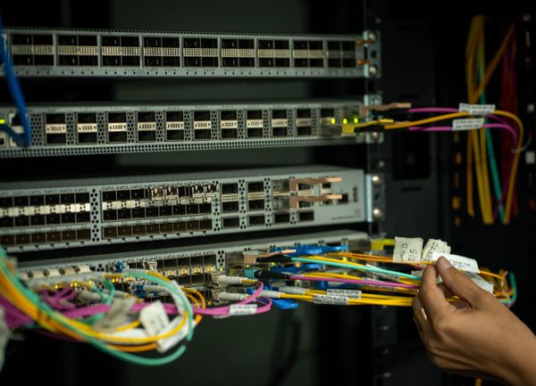 Mão humana ajustando cabos de fibra óptica coloridos conectados a servidores em um rack de data center.