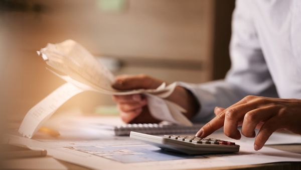 Pessoa utiliza calculadora e analisa documentos fiscais impressos sobre uma mesa de escritório.