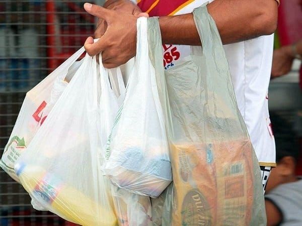 Pessoa carregando diversas sacolas plásticas brancas e verdes com produtos de supermercado.