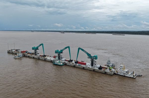 Píer flutuante com guindastes verdes operando no meio de um rio largo sob céu nublado.