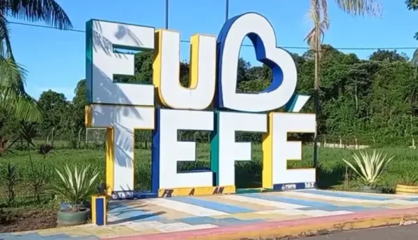 Letreiro turístico com os dizeres Eu Amo Tefé em cores azul, amarelo e branco, instalado em área aberta com vegetação.