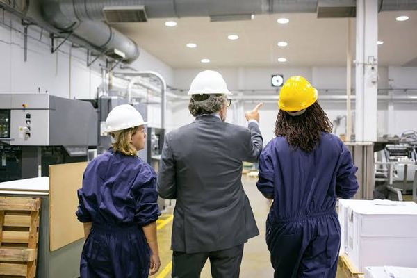 Três profissionais de costas, usando capacetes de segurança, observam o maquinário dentro de uma planta industrial.
