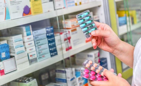 Pessoa segurando cartelas de comprimidos coloridos em frente a prateleiras repletas de caixas de medicamentos em uma farmácia.