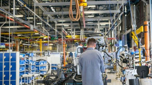 Operário de costas trabalhando em linha de montagem industrial com maquinários e estruturas metálicas ao fundo.