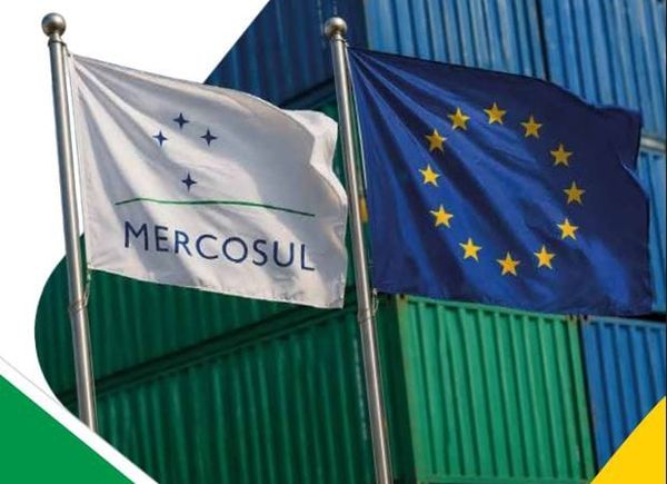 Bandeiras do Mercosul e da União Europeia hasteadas em frente a contêineres de carga empilhados.
