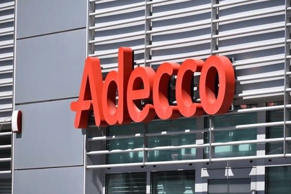 Logotipo da Adecco em letras vermelhas fixado na fachada de um edifício moderno com janelas de vidro.