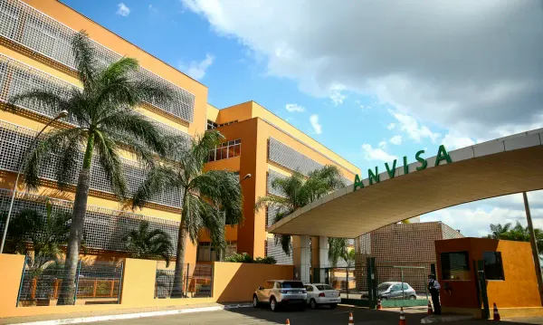 Fachada do edifício da Anvisa com palmeiras à frente e carros estacionados na entrada sob céu azul.