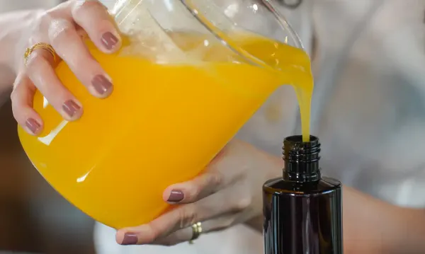 Pessoa despejando um líquido amarelo de uma jarra de vidro em um frasco escuro, em processo de fabricação de cosmético.