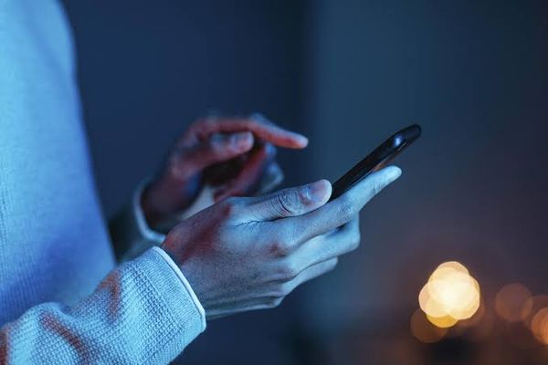 Pessoa segura smartphone com as duas mãos em ambiente com iluminação azulada e luzes desfocadas ao fundo.