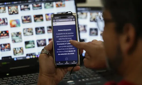 Pessoa segura celular exibindo aviso de acesso bloqueado a plataforma de apostas por determinação de Medida Provisória.
