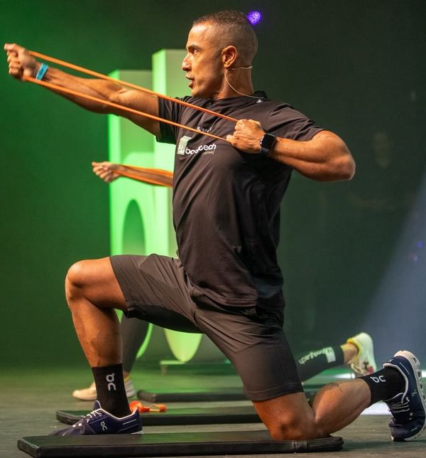 Homem ajoelhado em um colchonete realizando exercício com elástico extensor durante aula da academia Bodytech.