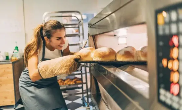 Mulher sorridente usando luvas térmicas retira uma bandeja com pães assados de um forno industrial em uma padaria.