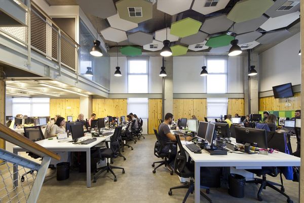 Colaboradores trabalhando em computadores em um escritório moderno com pé-direito alto e decoração industrial.