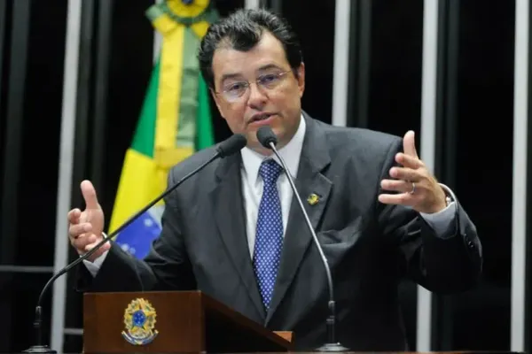 O senador Eduardo Braga discursa em um púlpito com microfones, vestindo terno escuro e gravata azul, com a bandeira do Brasil ao fundo.