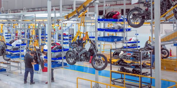 Linha de montagem da fábrica da Bajaj em Manaus, com motocicletas suspensas em suportes amarelos e funcionários trabalhando.